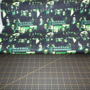 Puede incluir: Tela negra con un patrón repetido de imágenes de la película "Matrix Reloaded". Las imágenes incluyen personajes de la película, como Neo, Trinity y Morpheus, así como el título de la película.