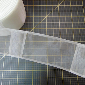 May include: A roll of white sheer ribbon with a rectangular design. The ribbon is made of a delicate, see-through material. The ribbon is on a black cutting mat with a grid pattern.