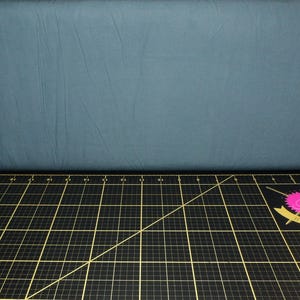 May include: A teal fabric backdrop above a black cutting mat with yellow grid lines. The cutting mat has measurements in inches. A pink and yellow logo is in the bottom right corner.