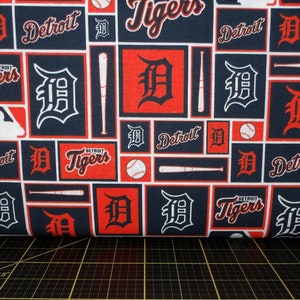 May include: A blue and red fabric with a repeating pattern of the Detroit Tigers logo, baseball bats, and baseballs. The fabric features the words "Detroit Tigers" and "Detroit" in a white font.