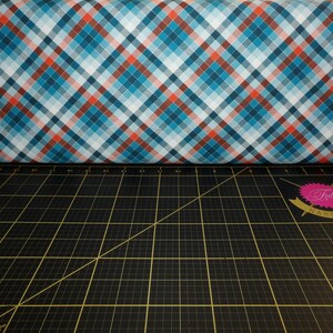 May include: A close-up of a blue, red, and white plaid fabric. The fabric is folded in half and is lying on a black cutting mat with a grid pattern.