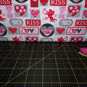 May include: A white fabric with a repeating pattern of red hearts, pink flowers, and the words "Kiss", "Love", and "XO".