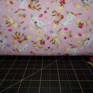 May include: Pink fabric with a repeating pattern of white bunnies holding carrots and baskets of Easter eggs. The background is a light pink with yellow, green, and blue accents.