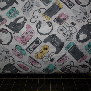 May include: A white fabric with a repeating pattern of various gaming controllers, headphones, and handheld gaming devices. The pattern is in shades of pink, blue, green, black, and gray.