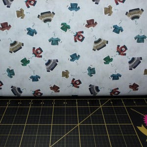 May include: Fabric with a repeating pattern of miniature sweaters in various colors, including red, green, blue, and brown, each hanging on a silver hanger. The sweaters are set against a light gray background.