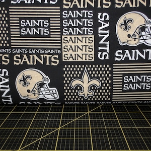 May include: Black fabric with a repeating pattern of gold Saints logos, including the fleur-de-lis and a football helmet. The text "Saints" is repeated throughout the fabric.