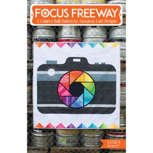May include: A quilt pattern featuring a camera with a colorful rainbow lens. The pattern is called "Focus Freeway" and is by Sassafras Lane Designs. The pattern number is #0057.
