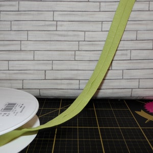 May include: A spool of lime green bias tape, unwinding onto a white plastic spool. The tape is lying on a black cutting mat with a grid pattern.