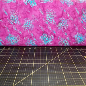 May include: Pink fabric with a repeating pattern of turquoise sea turtles. The fabric is on a black cutting mat with a ruler.