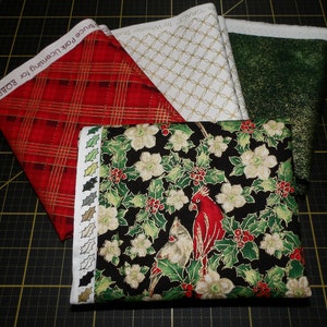 May include: Four fabric squares with a Christmas theme. The squares are red and gold plaid, white and gold geometric, green with gold glitter, and black with white flowers, red cardinals, and green holly.