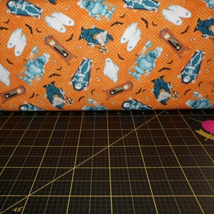 May include: Orange fabric with a repeating pattern of skeletons, ghosts, and clock faces. The fabric has a blue and white color scheme.