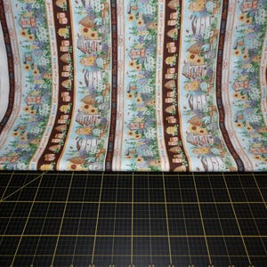 Puede incluir: Una tela blanca con un patrón repetitivo de tarros de miel, colmenas, girasoles y texto que dice "Bee Happy, Be Sweet, Be Kind, Be Yourself". La tela está doblada por la mitad y colocada sobre una estera de corte negra.