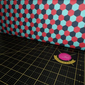 Puede incluir: Primer plano de una tela con un patr&oacute;n repetido de hex&aacute;gonos rojos, azules y blancos. La tela est&aacute; doblada sobre una estera de corte negra con un logotipo rosa y blanco que dice "Bella Pink Hardware".