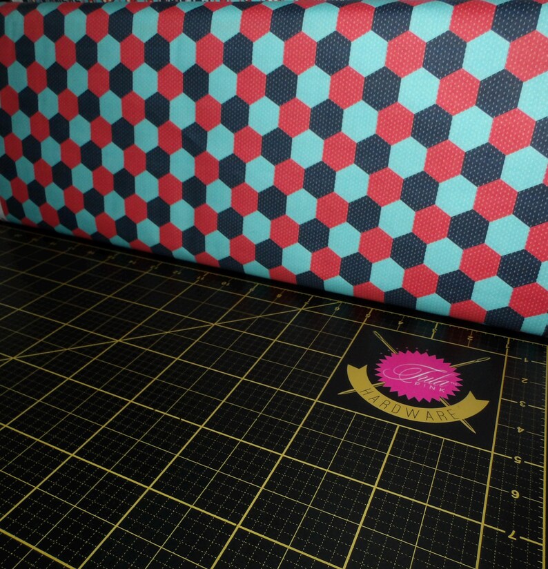 Puede incluir: Primer plano de una tela con un patr&oacute;n repetido de hex&aacute;gonos rojos, azules y blancos. La tela est&aacute; doblada sobre una estera de corte negra con un logotipo rosa y blanco que dice "Bella Pink Hardware".