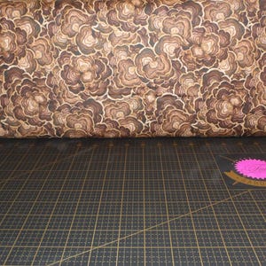 May include: Brown fabric with a repeating pattern of stylized flowers. The fabric is rolled up and sitting on a black cutting mat.