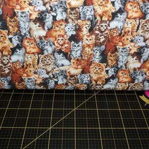 Puede incluir: Una tela blanca con un patrón repetido de muchos gatitos coloridos. Los gatitos están en varias poses y tienen diferentes colores de pelaje. La tela está sobre una estera de corte negra.