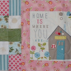 Puede incluir: Una colcha de retazos rosa y blanca con un diseño de casa en el centro. La casa es marrón con una puerta azul y un techo amarillo. El texto "Home is where you are..." está escrito encima de la casa.