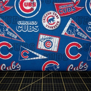 May include: Blue fabric with a repeating pattern of Chicago Cubs logos, including the team's name, a baseball, a pennant, and the team's logo with the letter "C" in a circle. The fabric is folded over a black and yellow cutting mat.