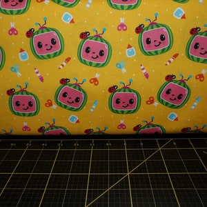 May include: Yellow fabric with a repeating pattern of cartoon watermelon TVs with ladybugs on top. The fabric is surrounded by a black cutting mat with white markings.