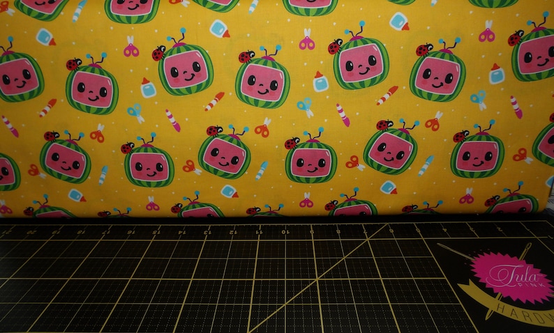 May include: Yellow fabric with a repeating pattern of cartoon watermelon TVs with ladybugs on top. The fabric is surrounded by a black cutting mat with white markings.