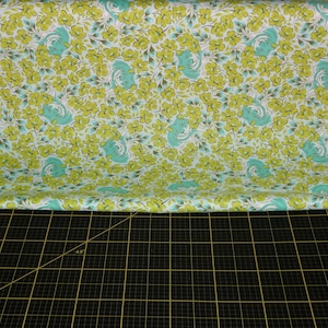 May include: A white fabric with a repeating pattern of green flowers and blue birds. The fabric is folded in half and laid on a black cutting mat.