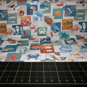 May include: Fabric with a repeating pattern of marine life illustrations. The design features squares with images of fish, starfish, crabs, seashells, and seahorses in shades of blue, orange, and beige. The fabric is folded.
