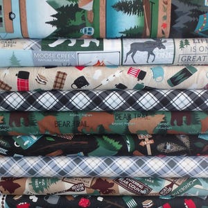May include: A stack of fabric featuring a camping theme. The fabric is printed with images of tents, campers, trees, and other camping-related items. The fabric is in a variety of colors, including green, blue, brown, and black.