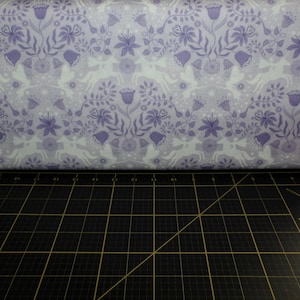 May include: Purple and white fabric with a repeating pattern of stylized flowers, leaves, and deer. The fabric is folded in half and lying on a black cutting mat.