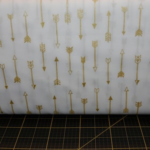 May include: White fabric with a repeating pattern of gold arrows. The arrows are pointing in different directions.