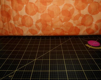May include: Orange fabric with a white background and a pattern of overlapping circles. The fabric is folded and lying on a black cutting mat with a grid pattern.