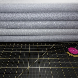 May include: Six rolls of patterned fabric in shades of grey, stacked on top of each other. The patterns include floral, dotted, and geometric designs. The fabrics are on a black cutting mat with grid lines.