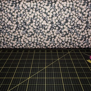 May include: A roll of fabric with a floral pattern. The fabric has a black background with small, light pink flowers and white stems. The fabric is resting on a black cutting mat with yellow grid lines.