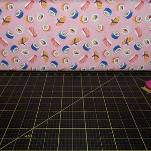 May include: Pink fabric with a repeating pattern of sushi rolls. The sushi rolls are in various colors, including pink, orange, yellow, green, and blue.