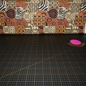 Può includere: Un rotolo di tessuto con un design patchwork. Il tessuto presenta motivi geometrici, strisce zebrate e motivi circolari nei toni del marrone, beige e nero. Poggia su un tappetino da taglio nero con linee a griglia dorate.