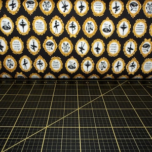 May include: Black fabric with a repeating pattern of gold frames containing images of ballerinas and swans. The fabric is on a black cutting mat.