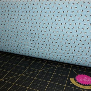 May include: A roll of light blue fabric with a repeating pattern of small bees in black and white. The fabric is resting on a black cutting mat with grid lines and a logo.