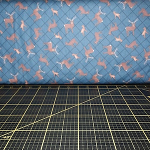 May include: A close-up of a blue fabric with a repeating pattern of brown deer. The fabric is lying on a black cutting mat with a white ruler and a pink label that says "Tula Pink".