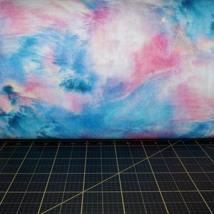 Może przedstawiać: Rolka materiału z wzorem w stylu akwareli. Wzór zawiera mieszane odcienie niebieskiego, różowego i białego, tworząc efekt chmury lub eteryczny. Materiał spoczywa na czarnej macie do cięcia.