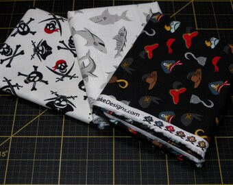 Puede incluir: Tres paneles de tela con diferentes diseños de temática pirata. El primer panel es blanco con calaveras y huesos cruzados negros, el segundo panel es blanco con tiburones grises y el tercer panel es negro con sombreros y ganchos de pirata rojos, blancos y grises. Los paneles están doblados y apilados uno encima del otro.