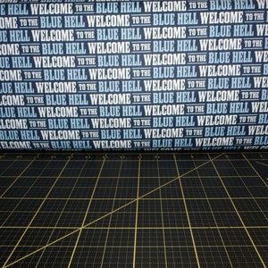 Peut inclure: Gros plan sur un tissu bleu et blanc avec le texte répété "Welcome to the Blue Hell". Le tissu est enroulé et partiellement visible sur un tapis de coupe noir.