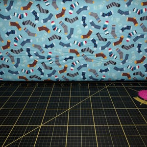 May include: A fabric roll featuring a repeating pattern of colorful socks in various designs. The socks are in shades of blue, brown, gray, and white, set against a light blue background. The fabric is likely cotton or a similar material, suitable for sewing projects.