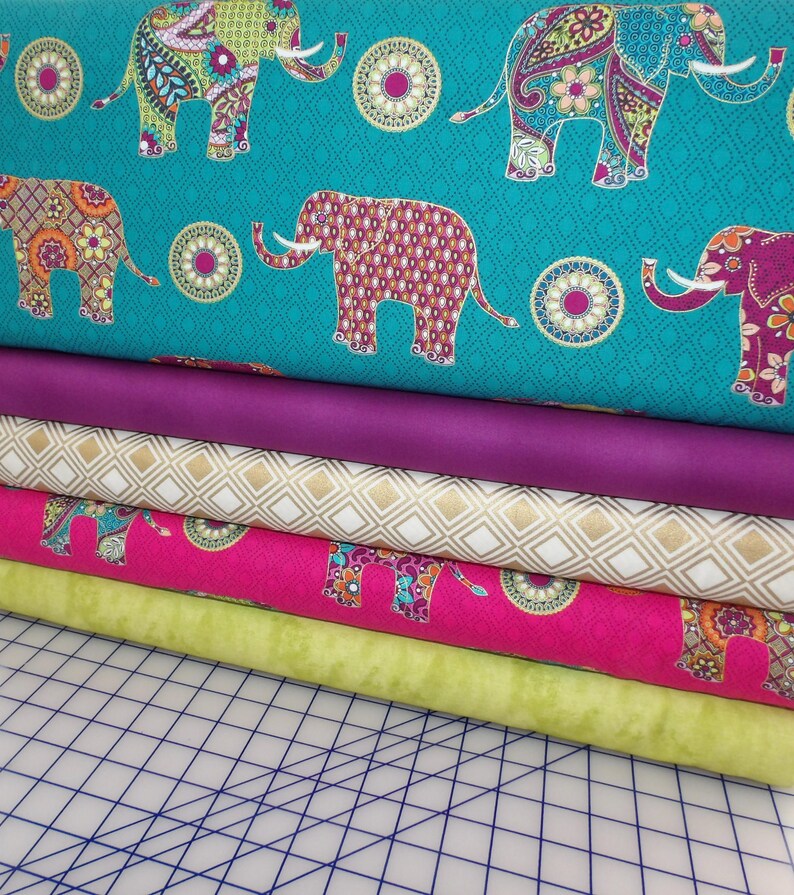 Puede incluir: Una pila de rollos de tela colorida. La tela superior es turquesa con elefantes estampados. Debajo hay telas lisas moradas, de diamantes dorados, rosas con estampado de elefantes y verde claro. Las telas est&aacute;n apiladas sobre una esterilla de corte con cuadr&iacute;cula.