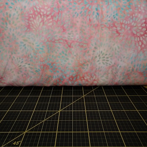 May include: Pink and blue batik fabric with a swirling pattern of white dots. The fabric is folded and lying on a black cutting mat.