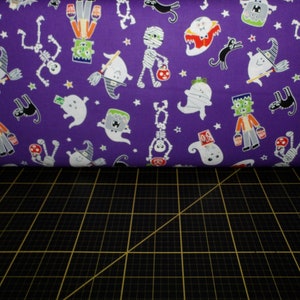 May include: Purple fabric with a Halloween print featuring ghosts, skeletons, mummies, witches, and monsters. The fabric is folded over a black cutting mat.