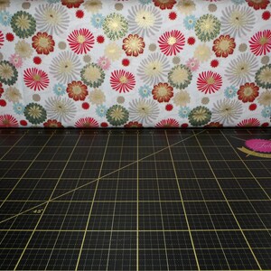 May include: White fabric with a repeating pattern of red, gold, and green stylised flowers. The fabric is folded in half and lying on a black cutting mat with a 45-degree angle line.
