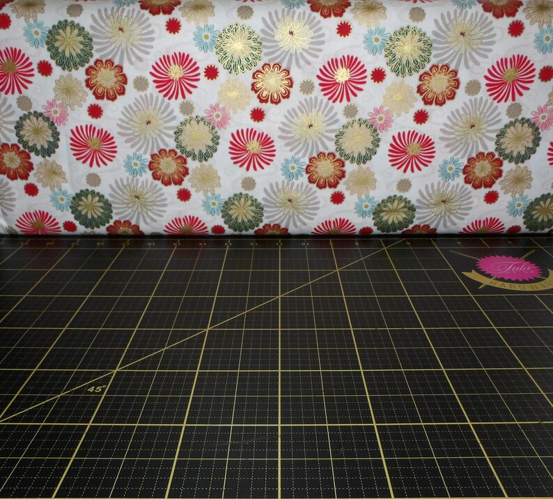 May include: White fabric with a repeating pattern of red, gold, and green stylised flowers. The fabric is folded in half and lying on a black cutting mat with a 45-degree angle line.