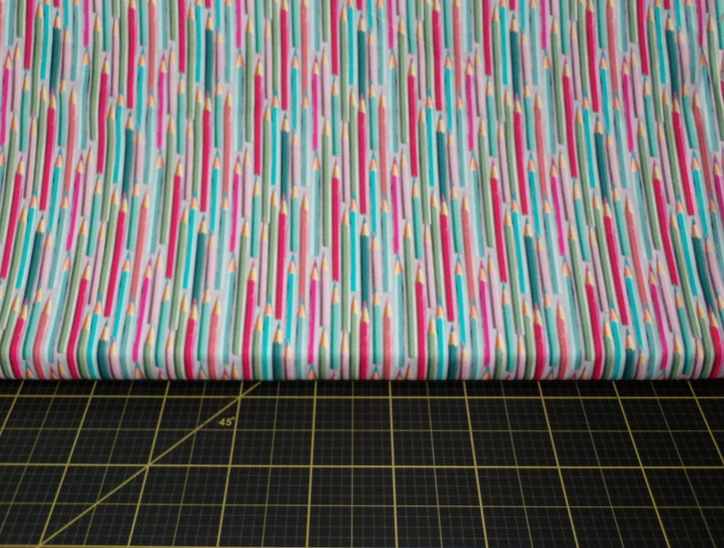 May include: A white fabric with a repeating pattern of colorful pencils. The pencils are in shades of pink, blue, green, and gold. The fabric is folded in half and placed on a black cutting mat.