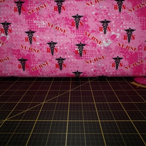 May include: Pink fabric with a repeating pattern of black medical caduceus symbols and the words "RN" and "NURSE" in red.