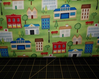 May include: A green fabric with a repeating pattern of colorful houses, trees, and fences. The houses are in various styles and colors, including red, blue, yellow, and green. There are also small trees and fences in the pattern.