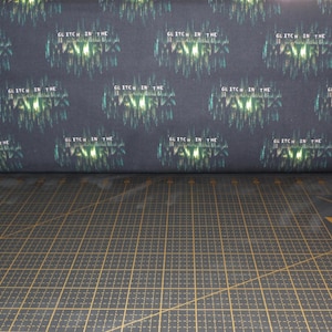 Puede incluir: Tela oscura con un patrón repetido de las palabras "GLITCH IN THE MATRIX" en un efecto de fallo verde y blanco. La tela está enrollada, mostrando el patrón. El fondo es una cuadrícula negra.